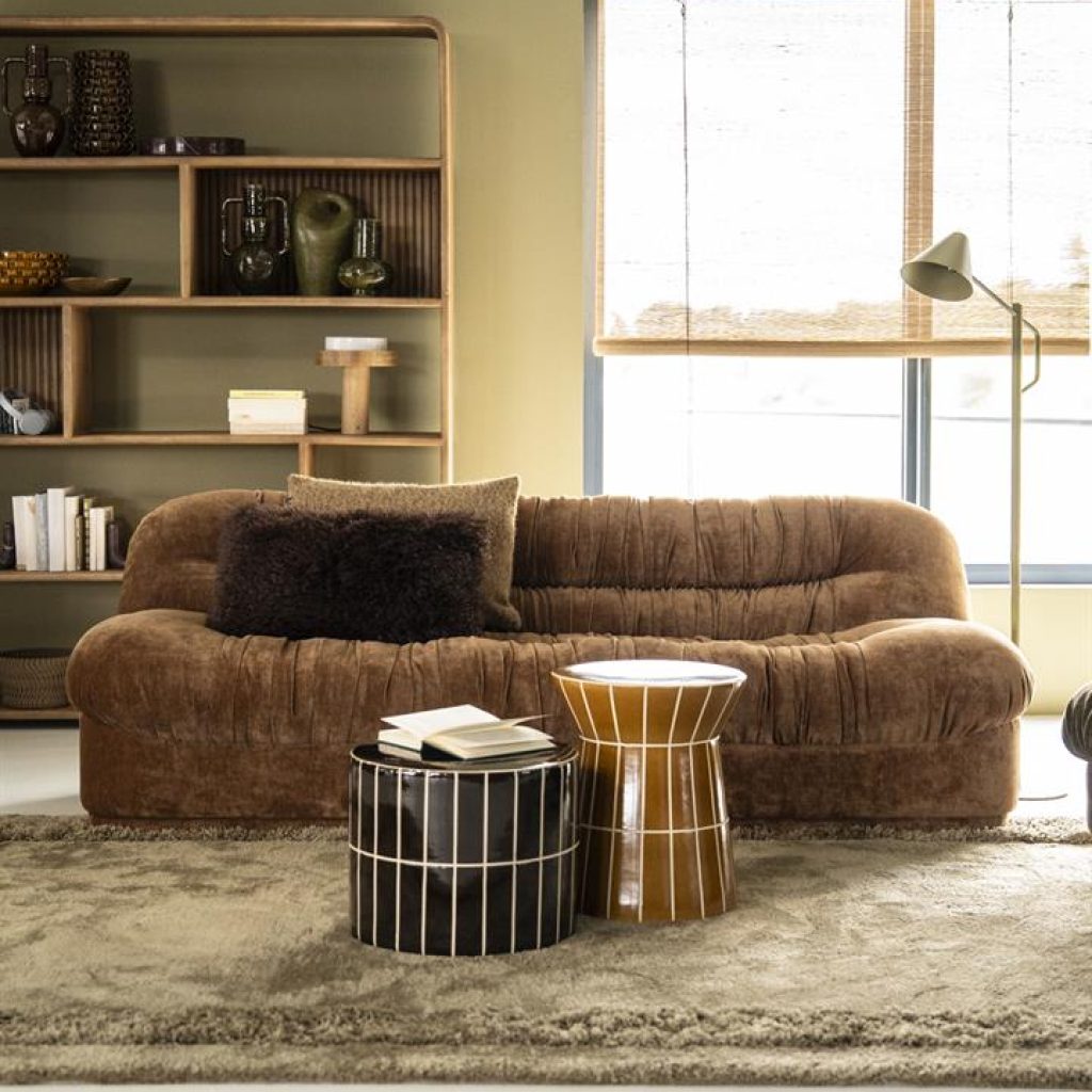 Een bruine retro bank in groene woonkamer, met groen vloerkleed en ronde tegeltjes bijzettafeltjes met houten vakkenkast achter de bank.