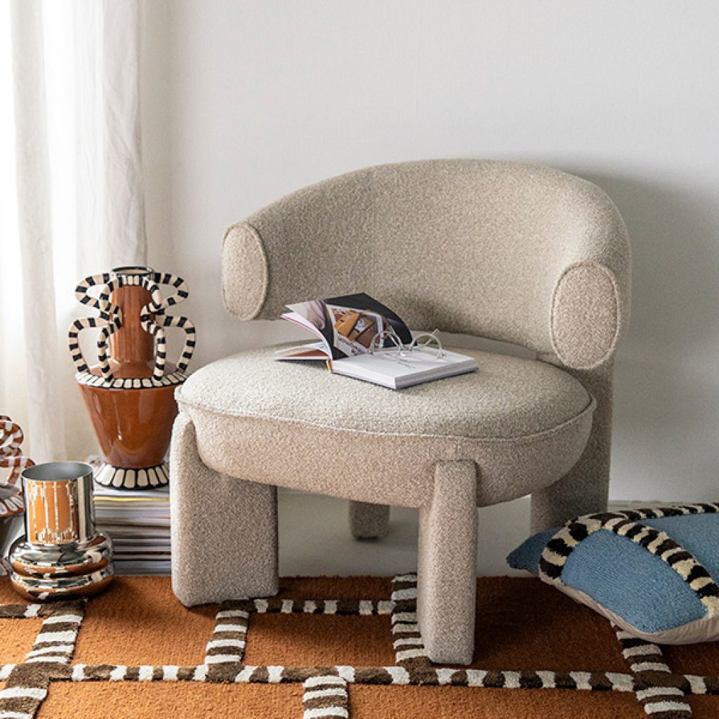 Beige fauteuil zonder armleuning, op bruin vloerkleed met print voor een witte muur.