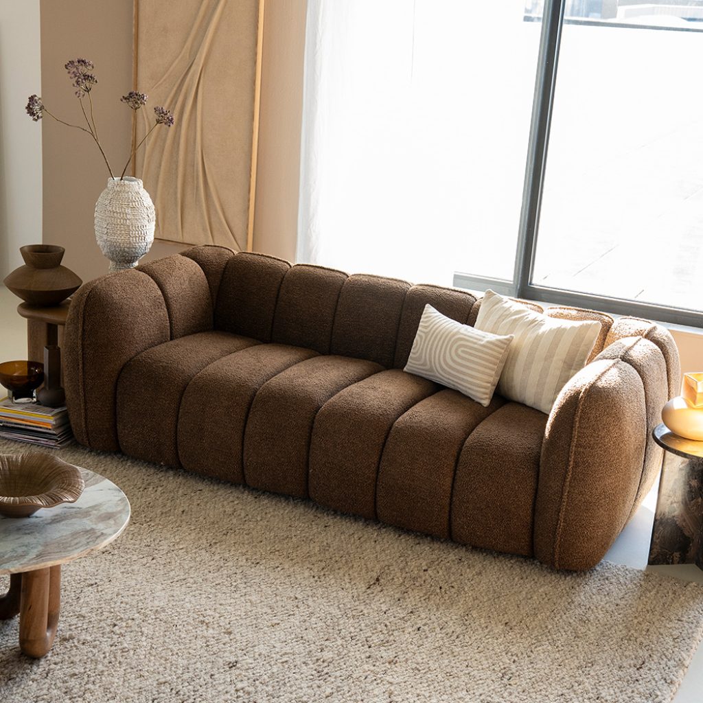Donkerbruine bank met verticale stiksels voor een beige vloerkleed in een natuurlijke setting met natuurlijke kleuren en materialen.