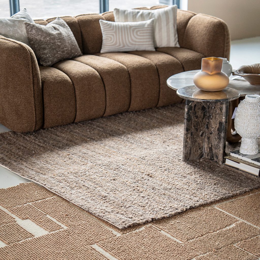 Een zithoek met rechte bruine bank met verticale stiksels, beige vloerkleden en bruin marmeren salontafel.