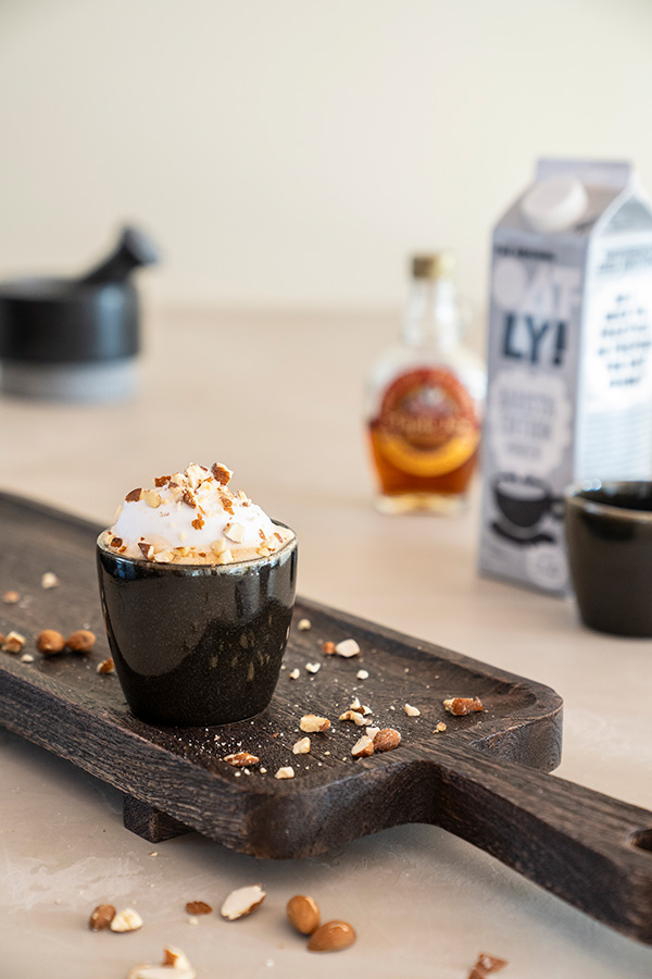 Sfeervol geserveerde cappuccino met toppings op een houten borrelplank, naast een pak haverdrank en siroop.